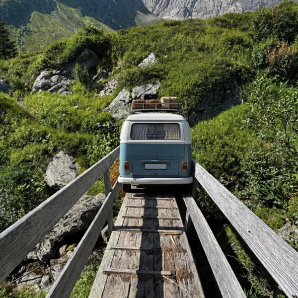 Ein alter Bulli fährt über eine schmale Holzbrücke in einer sommerlichen Berglandschaft